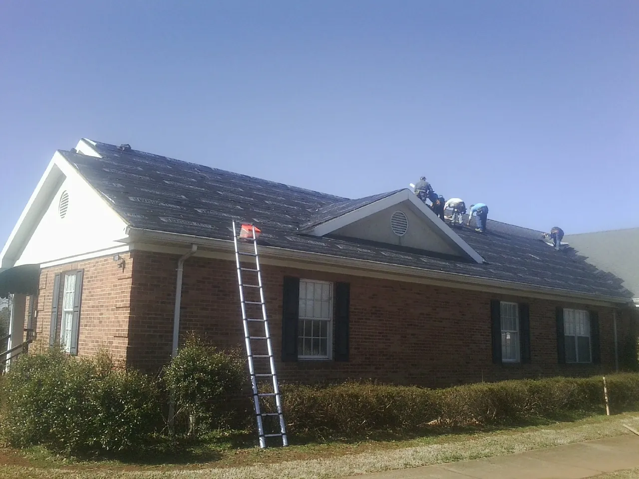 Crew tearing shingles off a brick ranch roof with a ladder set — Metal Roof Repair work by our crew in Conesville